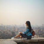 A young woman in casual attire sits on a rooftop terrace, viewing a sprawling cityscape in daylight.